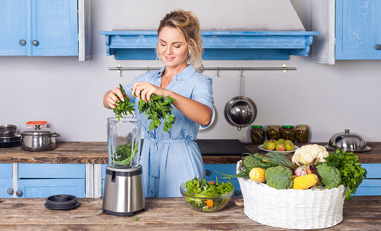 Woman in blue kitchen puts fresh herbs and leafy greens into a silver blender next to basket of fresh vegetables