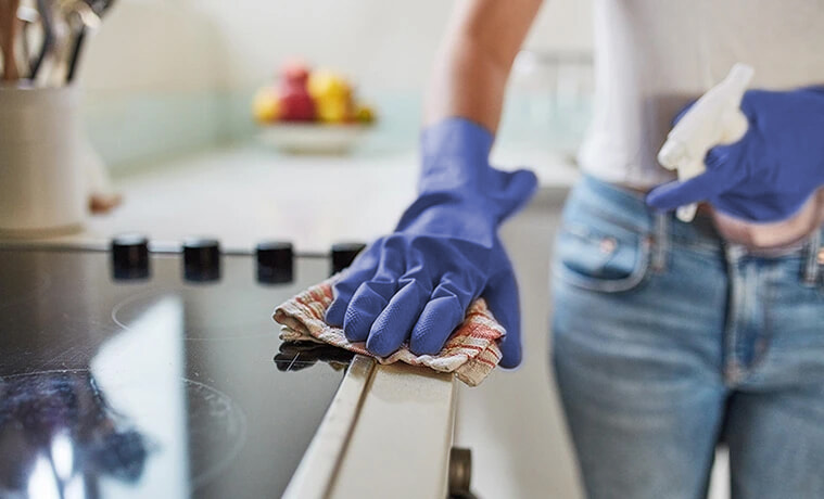 Woman wearing blue rubber gloves cleans her cooktop and kitchen bench with a cloth and cleaning solution
