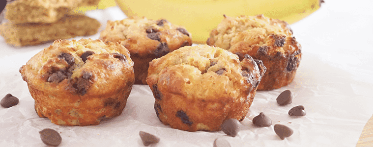 Banana and WeetBix Muffins on a white table with bananas and a scattering of chocolate chips