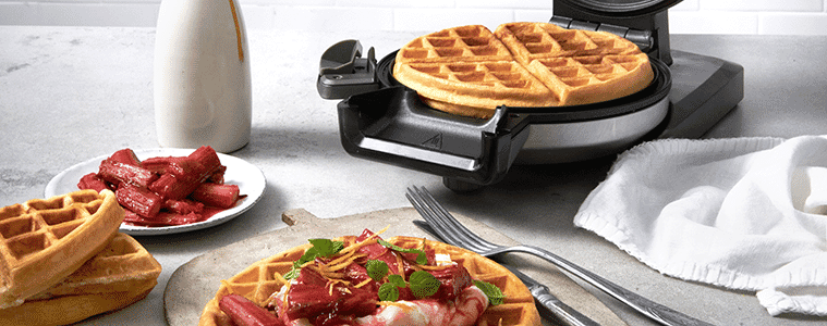 A waffle cooks inside a waffle maker a finished waffle sits on the table covered in toppings