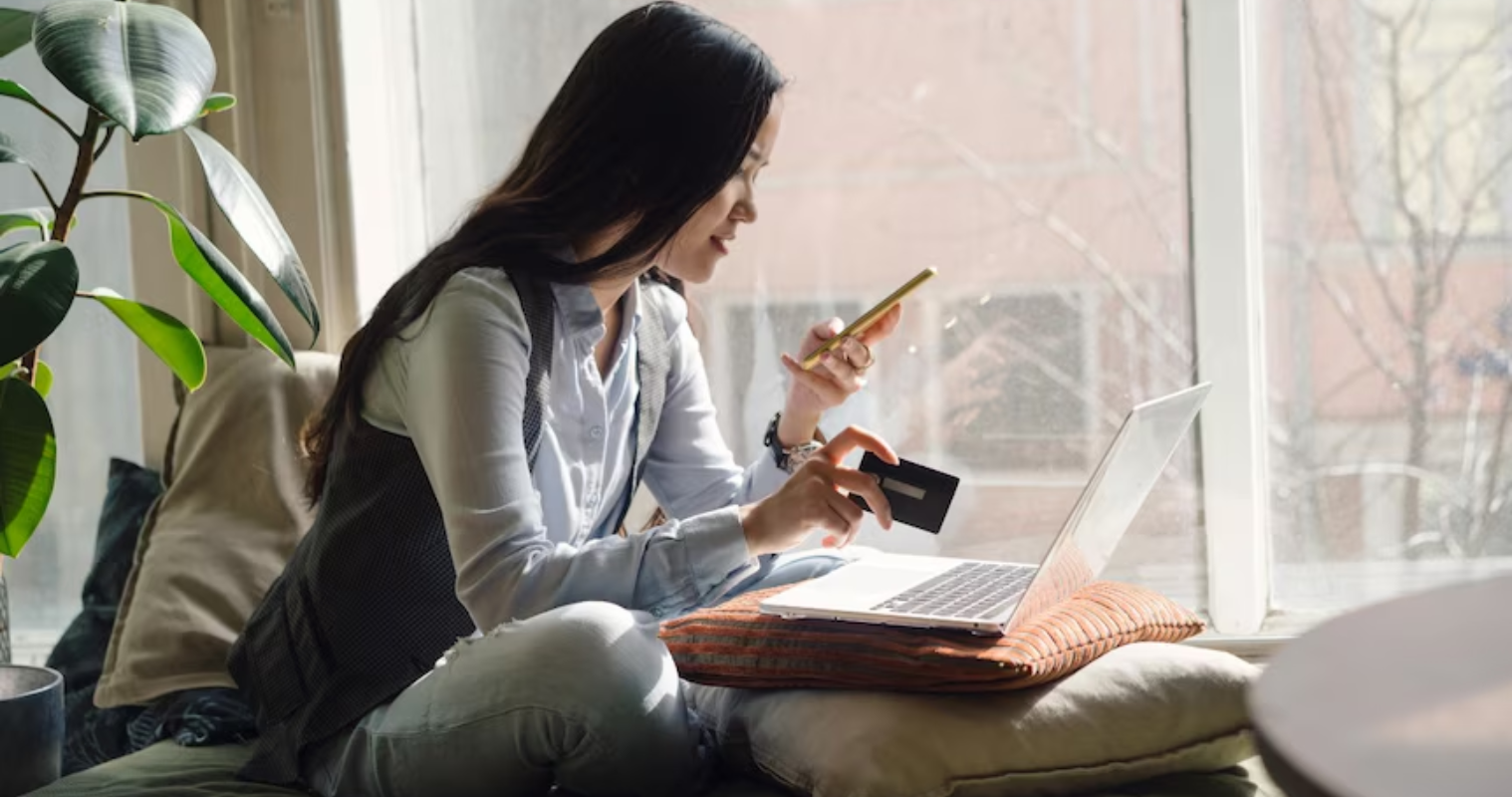 A woman sits on the bed credit card and smartphone in hand to do some online shopping with her laptop open in front of her