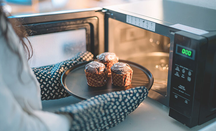 A woman with oven mitts pulling a tray of muffins out of a microwave