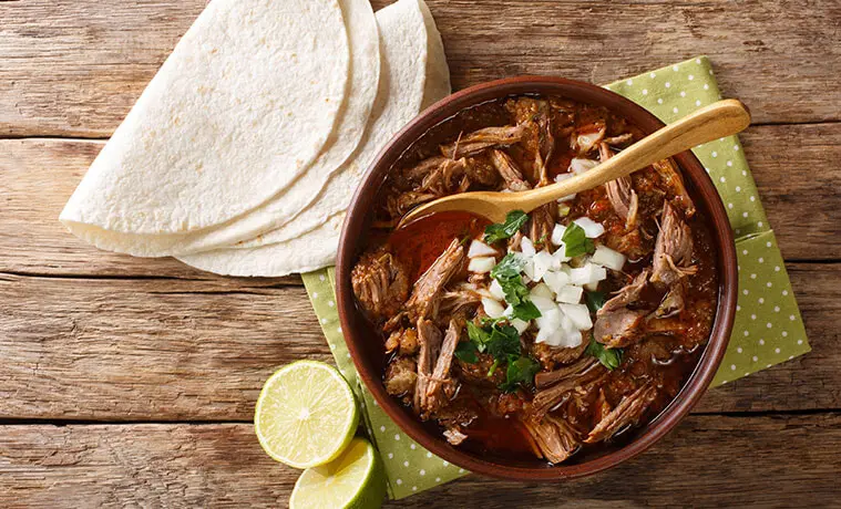 Soft tacos fanned on a timber table next to a ceramic bowl filled with slowcooked beef and a lime wedge