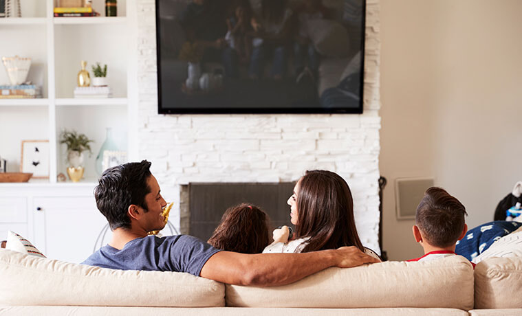 Back view of a young family of four sitting on sofa watching a mounted TV mum looking at dad
