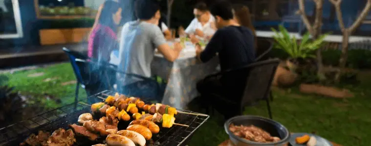 A closeup of meats cooking on a BBQ a family eats in the background
