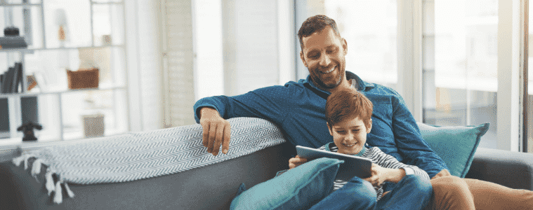 A young boy watches a video on a tablet while sitting with his father on the couch in their living room
