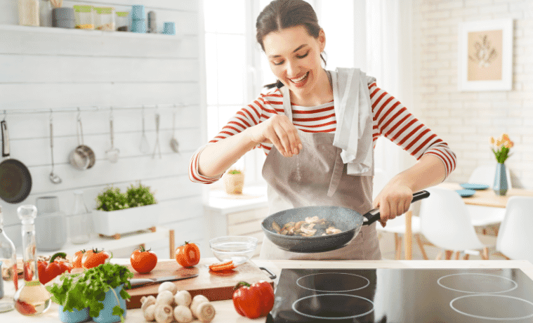A smiling woman seasons mushrooms in a frying pan she is holding above a sleek cooktop set into the kitchen bench