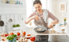 A smiling woman seasons mushrooms in a frying pan she is holding above a sleek cooktop set into the kitchen bench