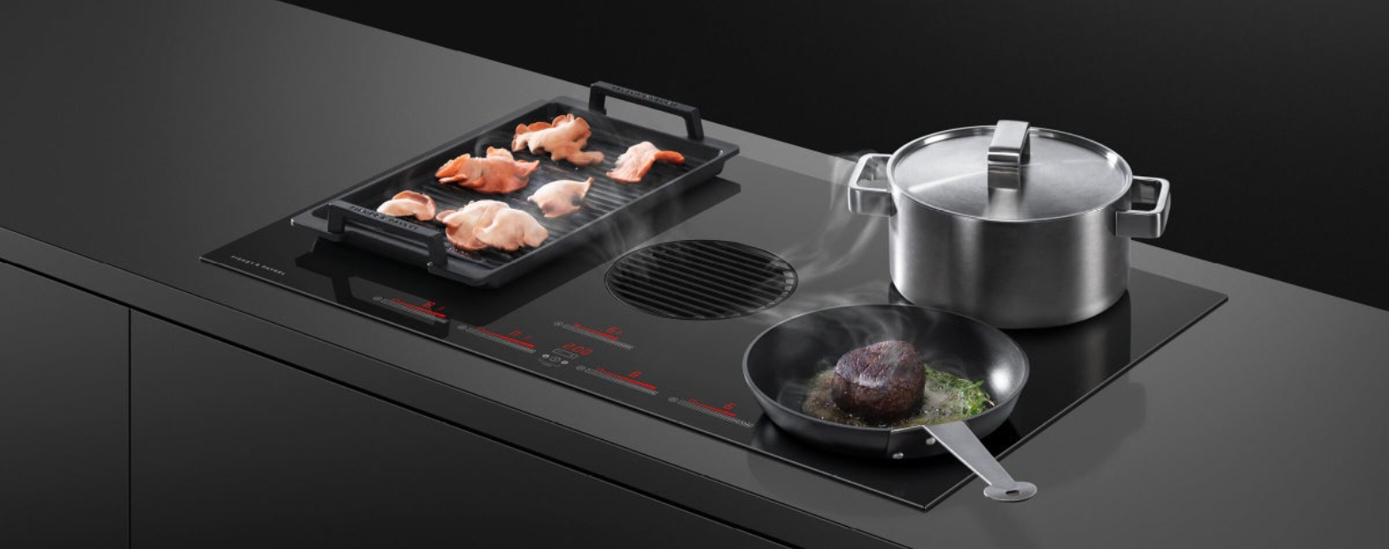Fisher & Paykel black induction cooktop with built-in downdraft ventilation, featuring a grill plate cooking bacon, a frypan searing steak, and a stainless steel pot on a dark kitchen island.