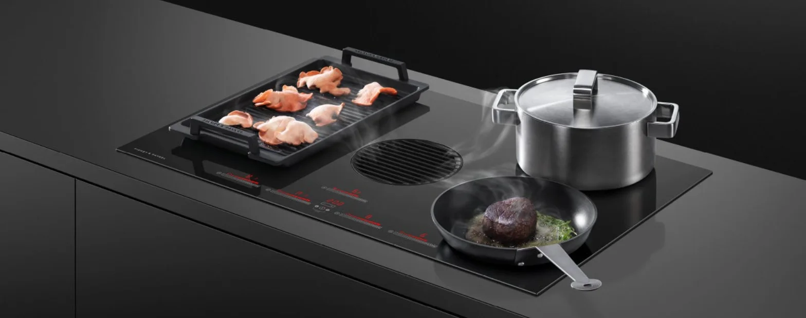 Fisher & Paykel black induction cooktop with built-in downdraft ventilation, featuring a grill plate cooking bacon, a frypan searing steak, and a stainless steel pot on a dark kitchen island.