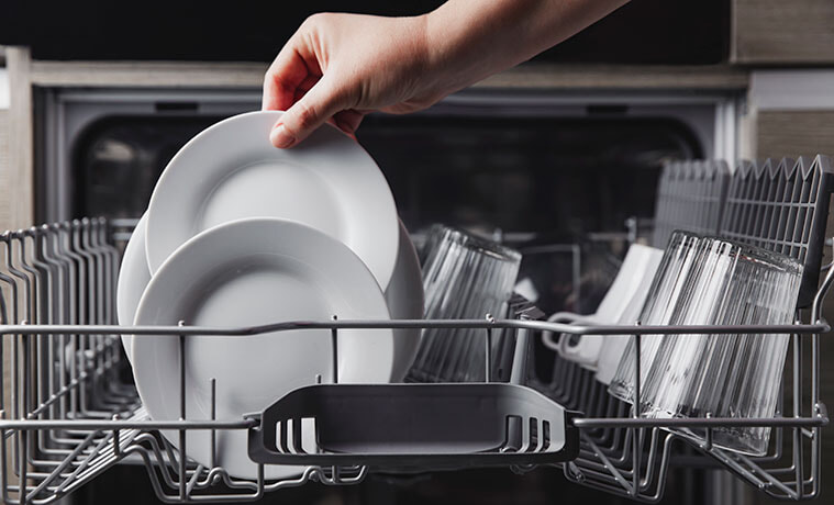 Closeup of a hand removing clean plates and glasses from the top rack in an open dishwasher