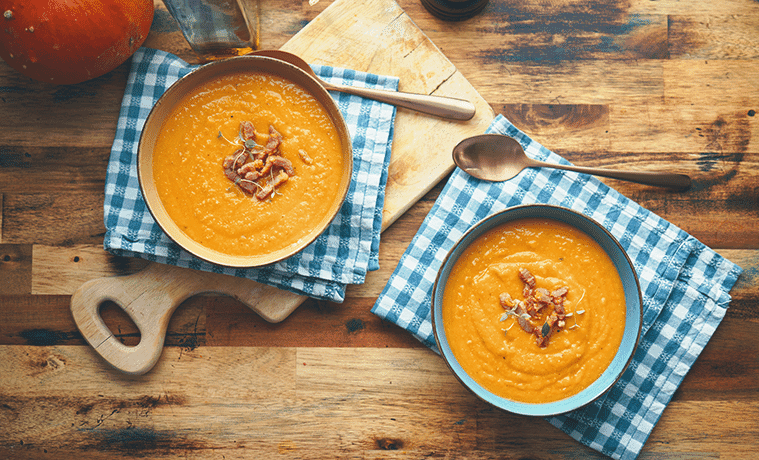 Two bowls of creamy vegetable soup sitting on blue gingham napkins on top of a timber cutting board and timber table