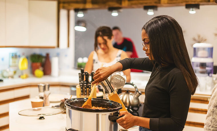 Young woman in a modern kitchen stirring a hot dish in a slow cooker with friends in the background helping with meal preparation