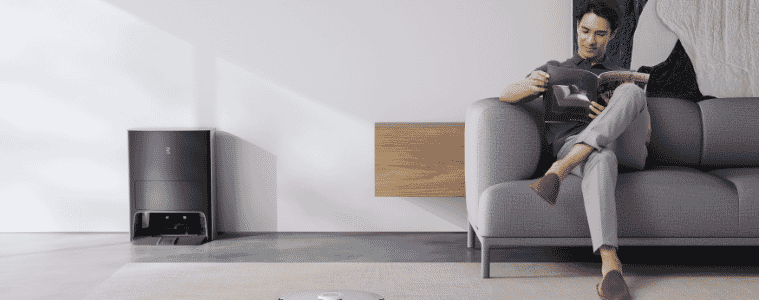 Man sitting on his couch casually reading whilst his robot vacuum cleans for him 