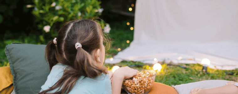 A young girl lies outside next to a bowl of popcorn watching a movie on a projector screen