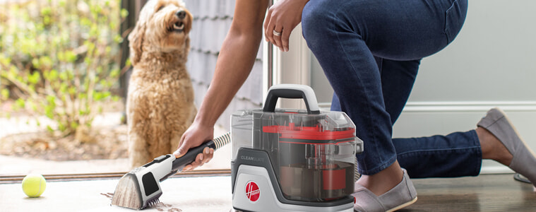 A woman uses her Hoover CleanSlate SpotWasher Carpet Cleaner to clean muddy pawprints off her entryway rug while her dog watches outside