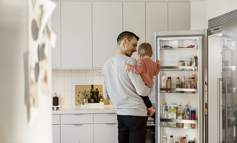 A father holds his toddler daughter while opening the door of his sidebyside fridge in a modern allwhite kitchen