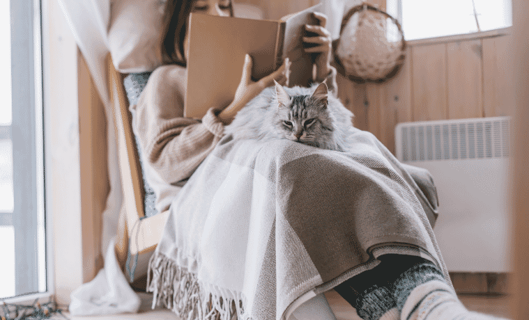 a woman sitting by her heater reading a book with a cat on her lap 