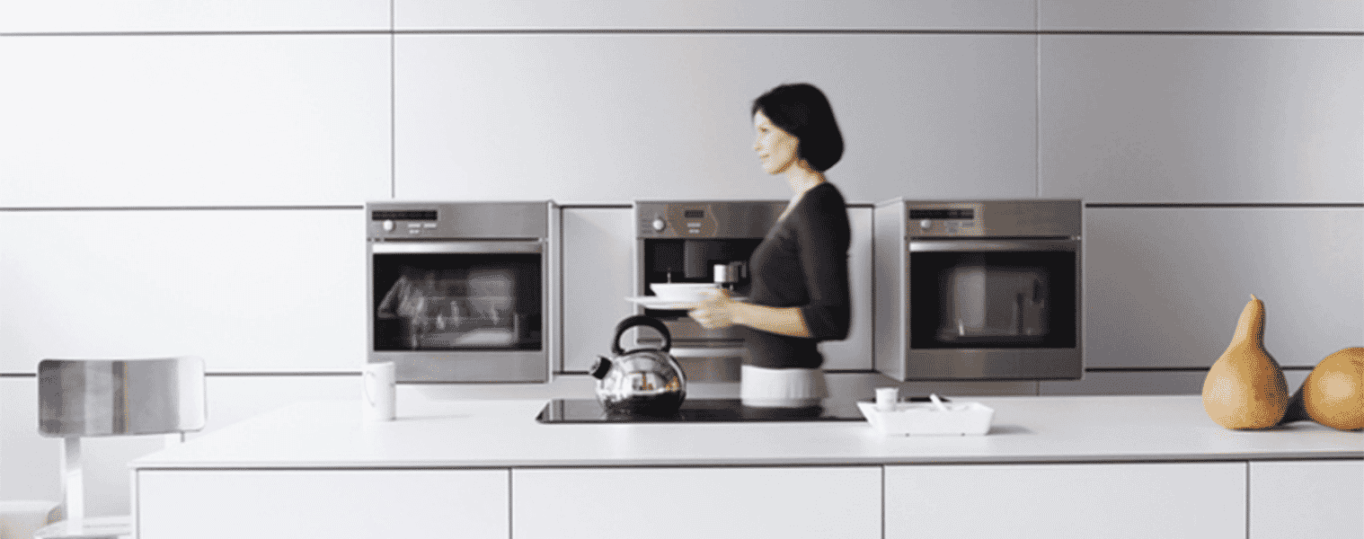 Woman walking through modern light coloured kitchen past a bank of ovens with pyrolytic cleaning