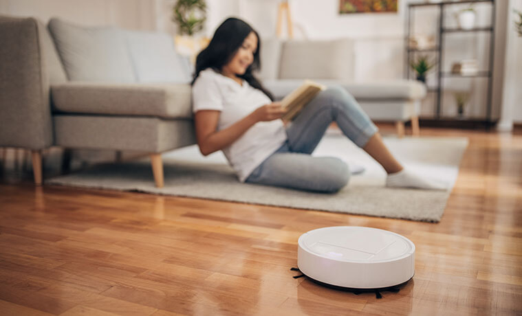 A woman reads a book on the floor of her living room while a robot vacuum cleans the timber floor