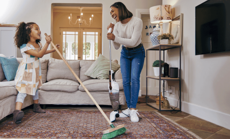 A mother and daughter have fun cleaning the living room with a stick vac and a broom
