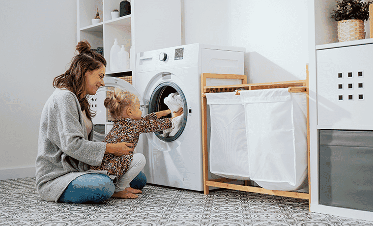 A mother and daughter sit on the laundry room floor to place clothes in the front loader washing machine