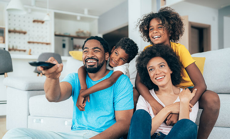 A smiling father holds a black remote and enjoys family viewing time with his wife and children