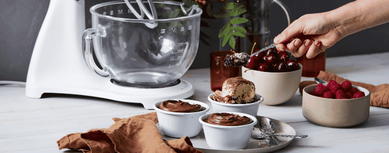 A Smeg stand mixer beside ramekins of individual Nutella puddings fruit and chocolate sauce in a grey background and a hand holding a serving spoon