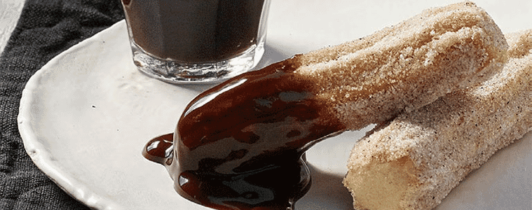 Two churros dipped in chocolate on a white plate sitting on a black napkin next to a cup of black coffee
