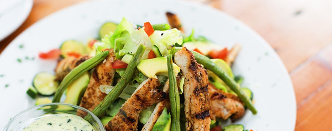 Healthy grilled chicken salad on a white plate with a bowl of dressing on the side 