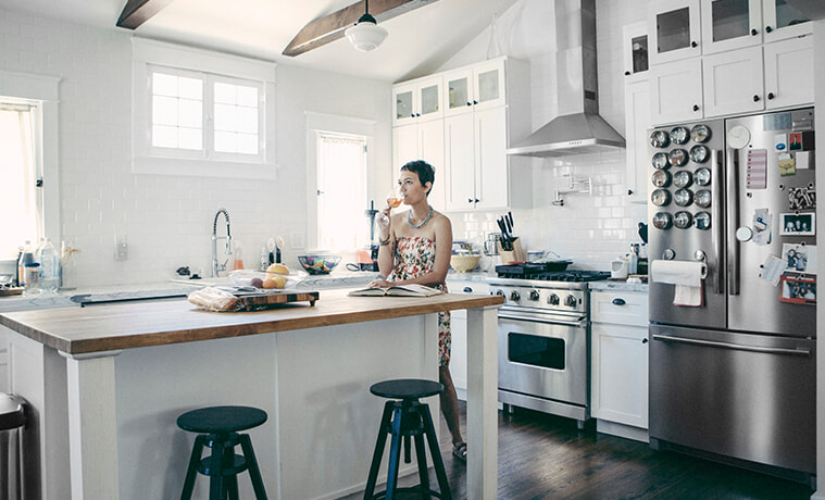 A woman drinks a glass of wine while looking at a cookbook in her kitchen