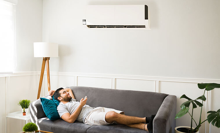 A man lies on the couch in his living room and turns on the wallmounted air conditioner using a remote control