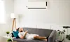 A man lies on the couch in his living room and turns on the wallmounted air conditioner using a remote control