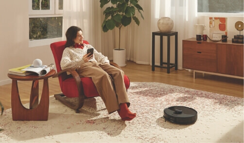 Image of woman smiling in a living room chair as a Roborock robotic vacuum works on a rug