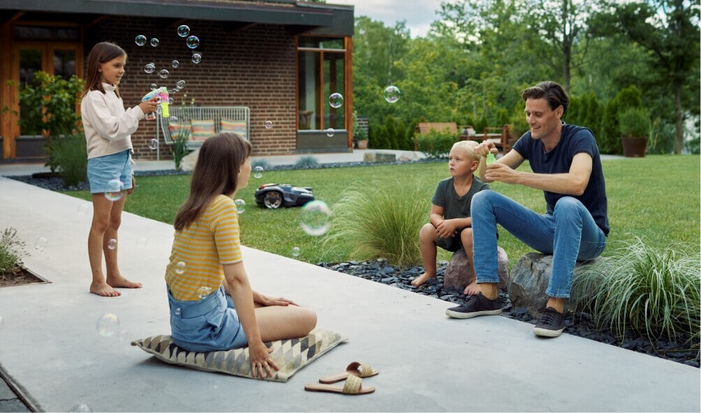 A father and his three children blow bubbles while a Husqvarna robot lawn mower mows their lawn
