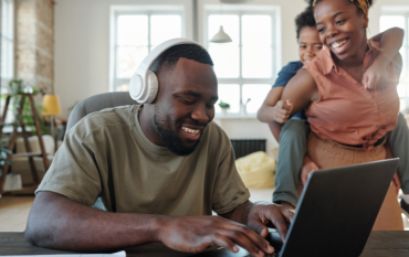 A smiling dad wearing white headphones at a laptop, with his family behind him