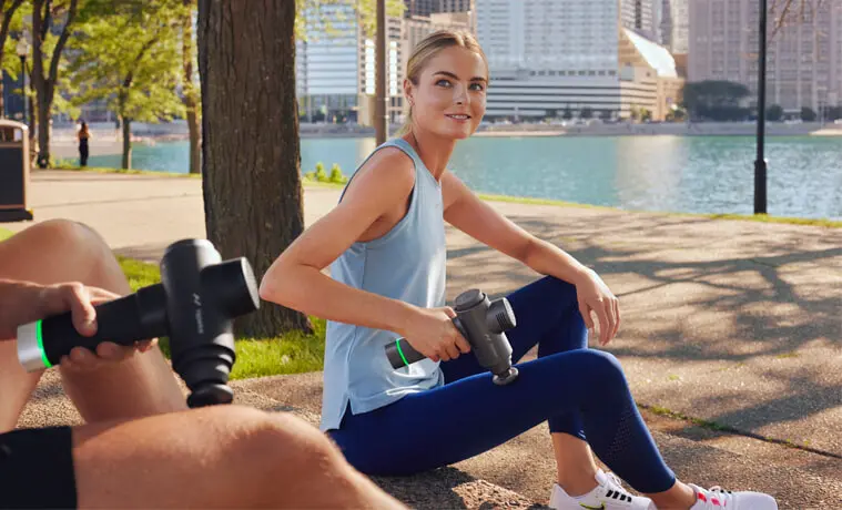 A woman in a park uses the Hyperice Hypervolt massage gun on her leg in a park