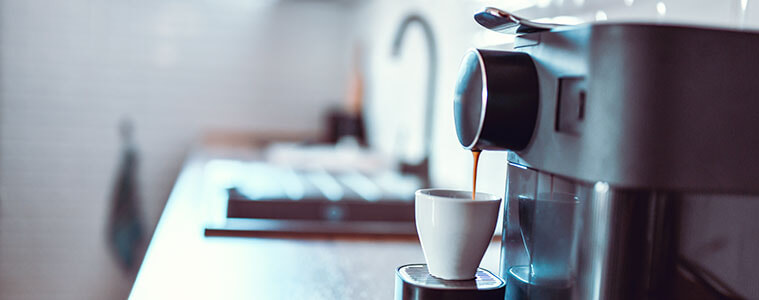 Woman holding a mug of fresh coffee looks at her coffee machine on the kitchen bench 