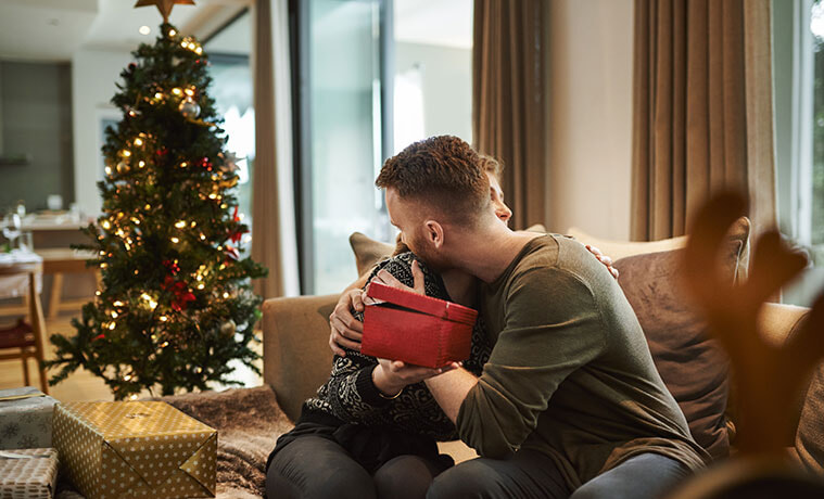 A young couple sitting on a couch near a Christmas tree embrace as they exchange gifts