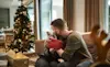 A young couple sitting on a couch near a Christmas tree embrace as they exchange gifts