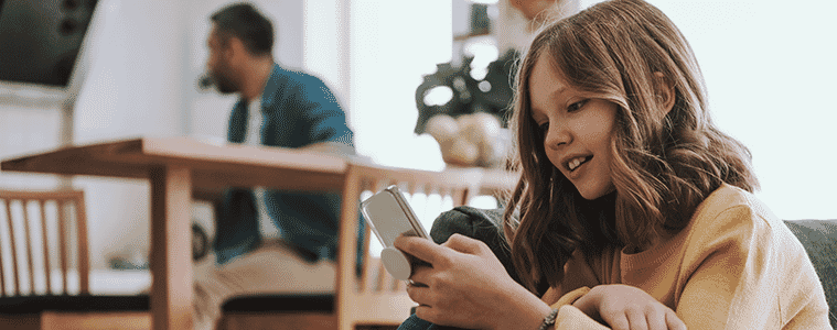 Young girl sits on a sofa busy with a mobile phone while father is on a video call at the dining table in the background