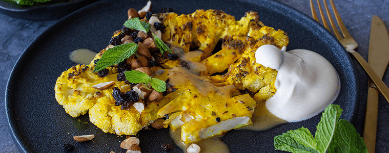 Cauliflower sliced and grilled covered with nuts green mint and currants with a dollop of yoghurt on a black plate