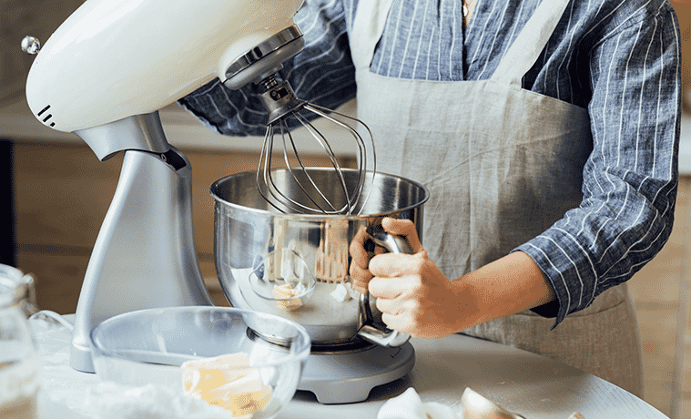 A woman wearing an apron lifts the whisk on her kitchen stand mixer and holds its bowl in her other hand 