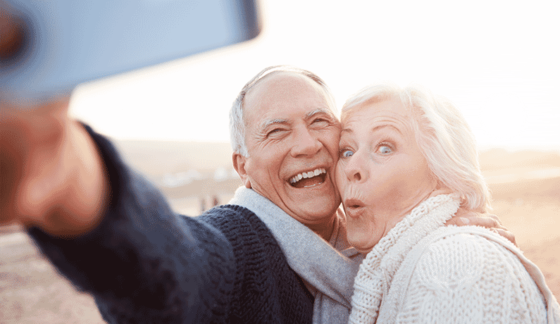 Lovely older couple are taking a selfie on the beach with their mobile phone The husband is smiling and the wife is making a funny face
