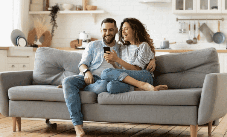 Woman and man snuggle on a couch laughing while looking at something on the womans smartphone screen