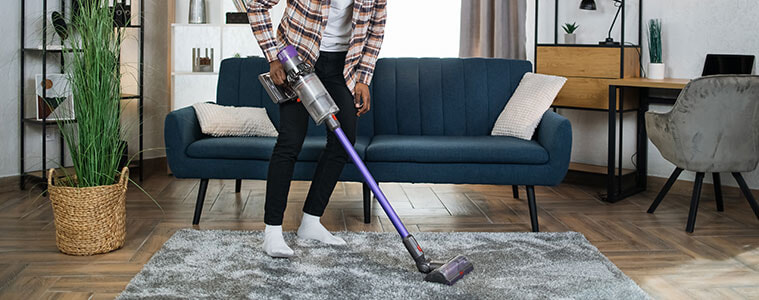 A young man wearing headphones listens to music while cleaning the living room floor with a stick vac