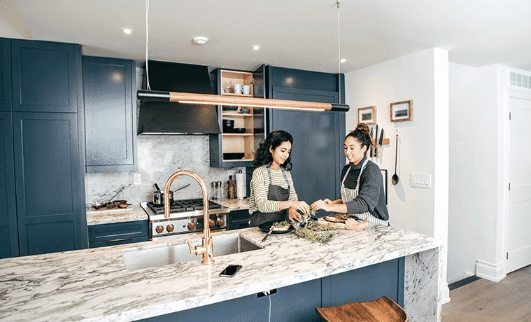 Two friends prepare dinner in a highend kitchen with marble benchtops blue cabinetry and brass taps