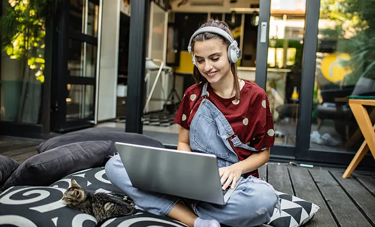 A teenage girl is at home wearing headphones and using her laptop while sitting on the back deck with her cat