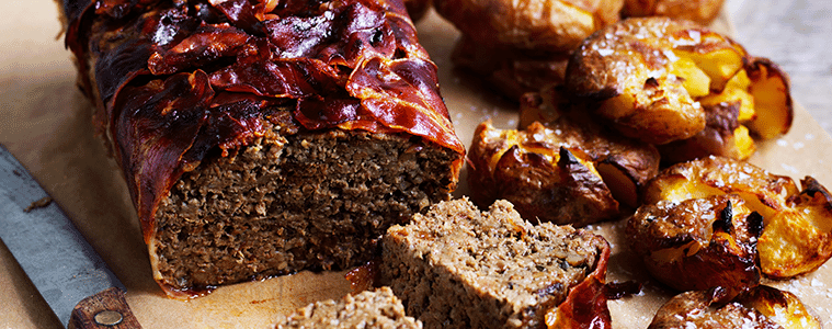 Meatloaf wrapped in bacon on a timber board a delicious slice in the foreground and roasted potatoes on the side 