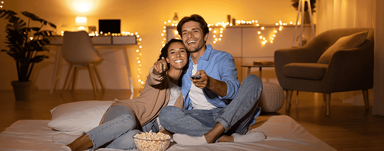 Couple sitting on their bed watching TV surrounded by fairy lights